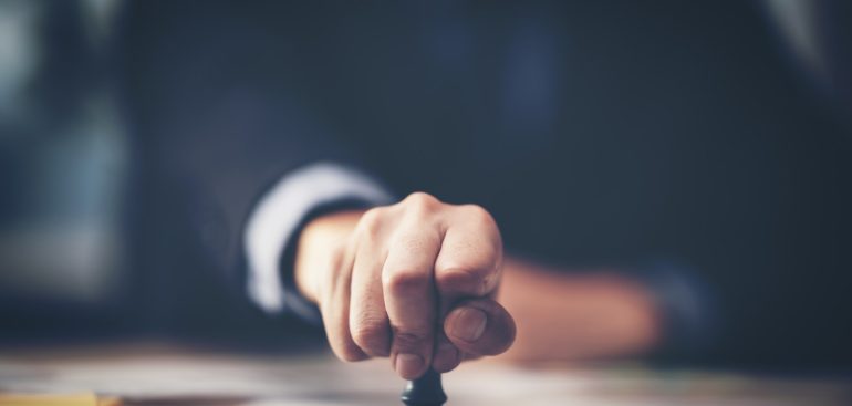 Close-up Of A Person's Hand Stamping With Approved Stamp On Document At Desk